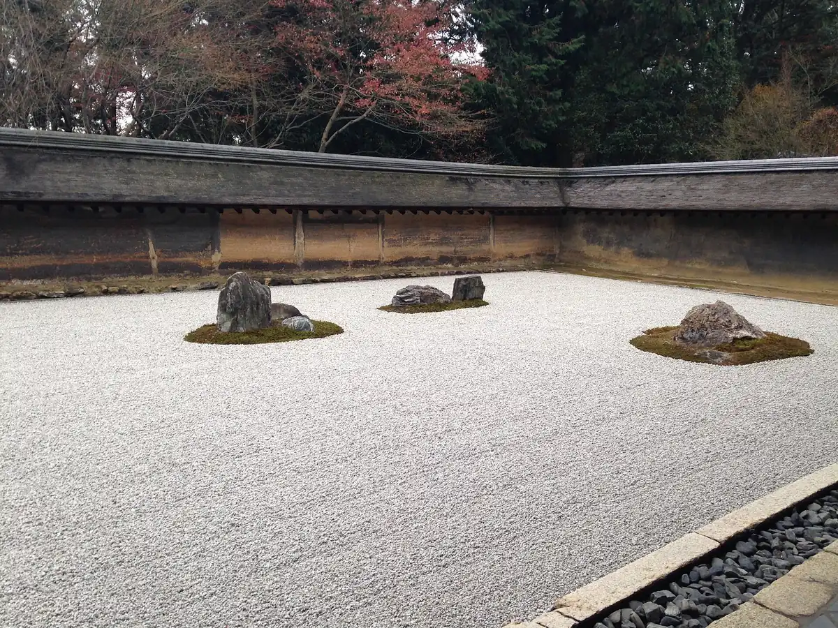 A dry rock garden with pale raked gravel and three clusters of stones on moss patches, set against an old wall with foliage behind it