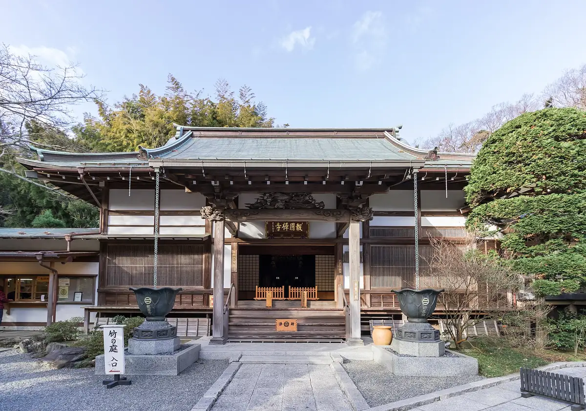 A wooden temple hall with a green copper roof, stone steps and a bronze urn on each side