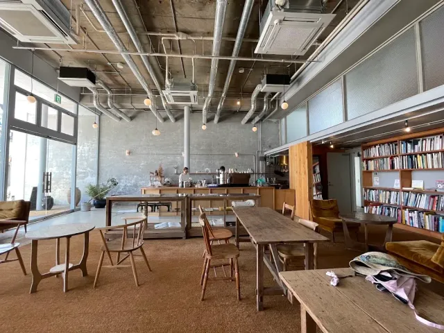 A concrete space with exposed pipes, heavy wooden tables and a wall of books