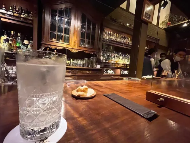 A glass of a sparkling drink on a wooden counter, with a bottle cabinet and bartenders behind
