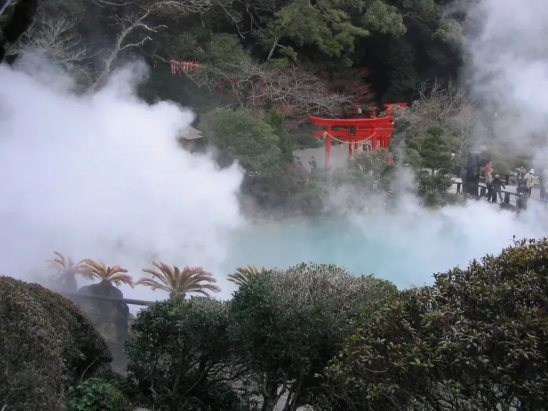 White steam rising from a geothermal pool with a red torii gate on the slope above it