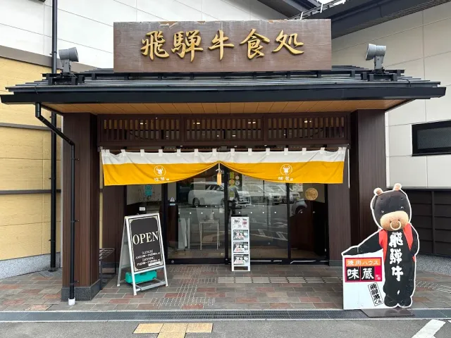 A wooden frontage with a carved sign, a white and yellow curtain and a smiling cardboard figure by the entrance