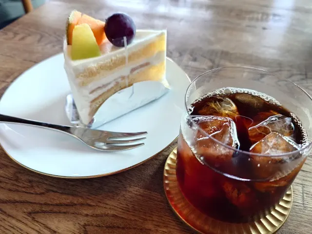 A slice of layer cake with fruit on a white plate, beside a glass of iced coffee