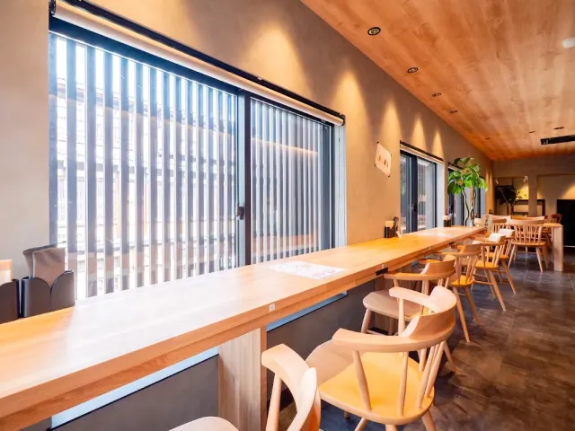 A light wooden counter along a large window with wooden chairs and a panelled ceiling