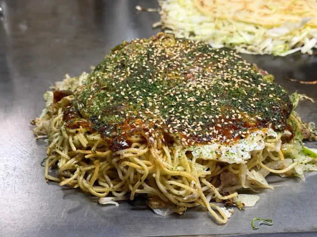 A savory pancake on a hot metal griddle, covered with noodles, brown sauce and sesame