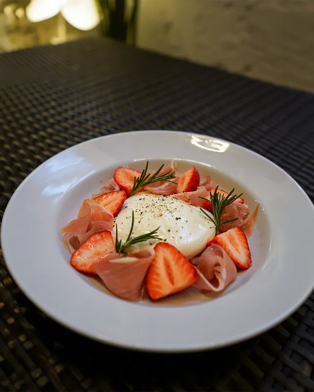 A round white cheese in a deep plate, surrounded by strawberries, cured ham slices and rosemary