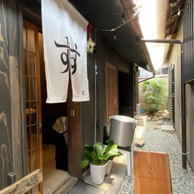 An entrance in a narrow alley with a white curtain and a Japanese character, a dark wooden building and a green plant