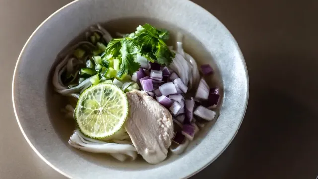 A grey bowl with noodles in broth, coriander, chopped red onion, a lime slice and a piece of chicken