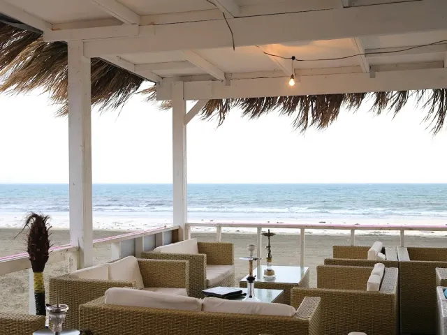 A terrace roofed with straw holding wicker sofas and tables, facing a beach and grey sea