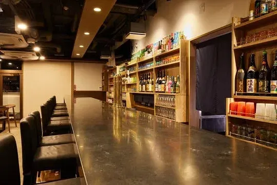 A long polished counter with black stools, and wooden shelves loaded with bottles