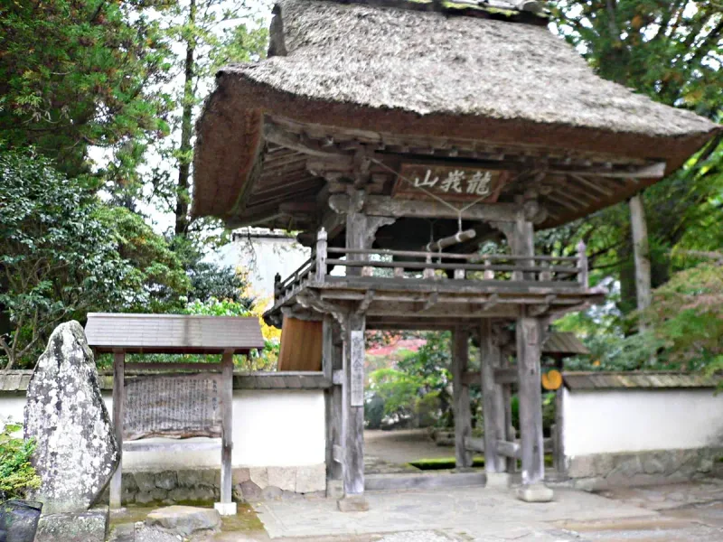 A wooden temple gate with a thick thatched roof, stone markers and green trees around it