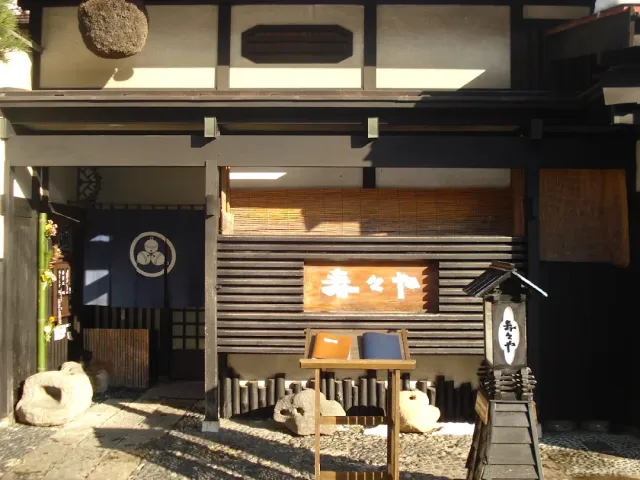 A traditional dark wooden frontage with a carved sign, a hanging ball of branches and a low bamboo fence