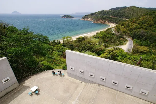 A concrete building on a green slope above a bay and beach