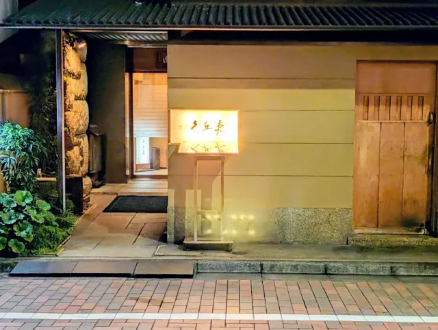 An entrance at night with a small lit sign, a stone wall, a wooden door and a tiled eave