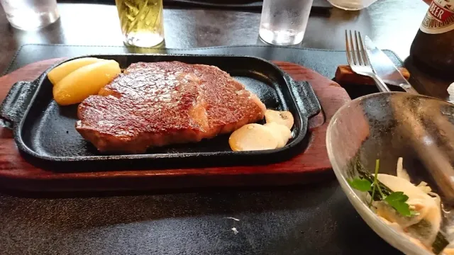 A grilled beef steak on a cast iron pan beside potatoes, with a salad bowl at the side