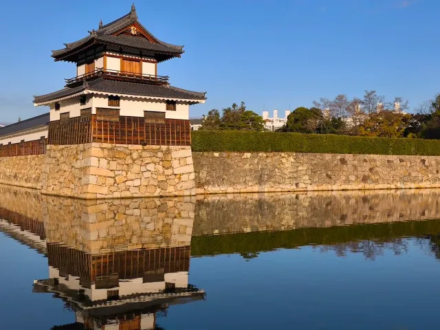 A wooden turret on a stone wall reflected in moat water, under a clear sky