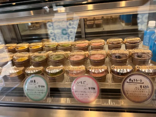 A display fridge with rows of dessert cups in different flavors and handwritten price signs