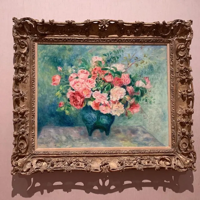 An oil painting of a bouquet of roses in a vase, in a carved gilded frame on a pink wall