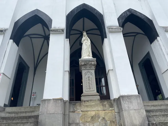 A white statue of Mary on a stone pedestal, in front of pointed arches on a white facade