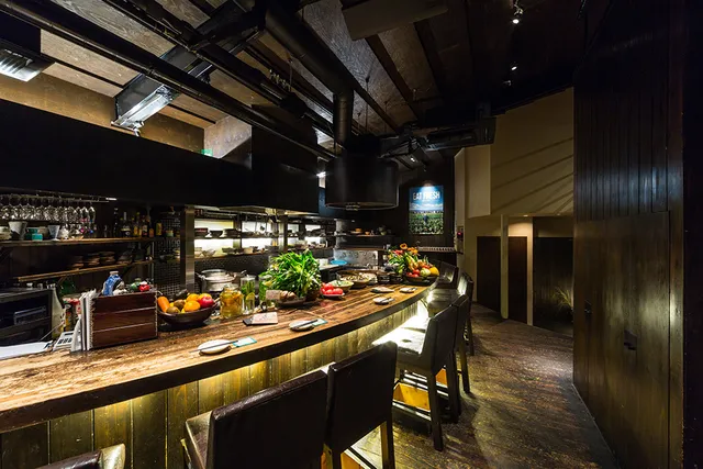 A long wooden counter with fresh vegetables and high chairs facing an open kitchen
