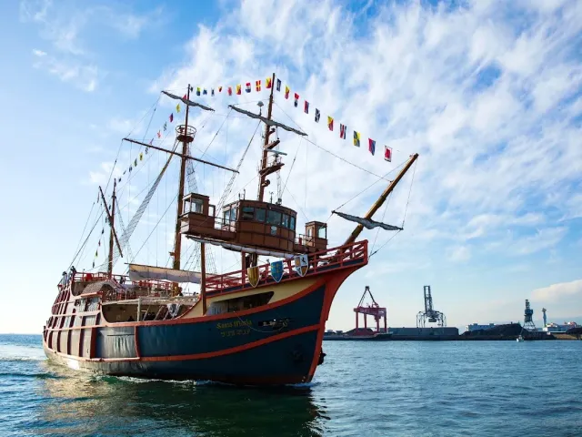 An old style sailing ship with colorful pennants, sailing on the water near harbor cranes