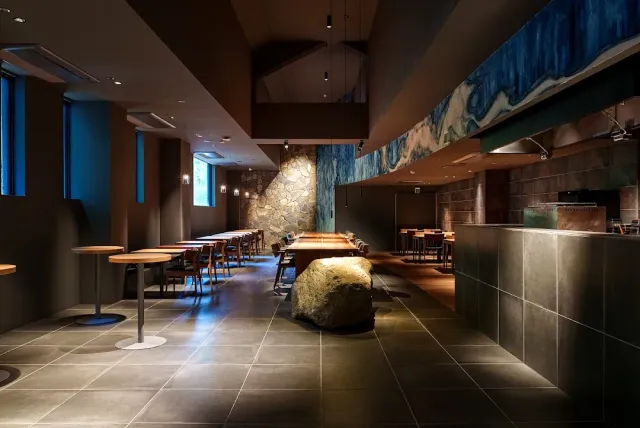 A dim space with a counter, wooden tables, a large boulder on the floor and a wall painted blue